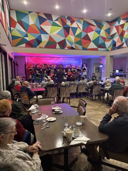 The Chamber Chorale of Frederickburg singing on an elevated stage in a dining room with tables and chairs full of senior citizens singing. The room is decorated with geometric pattern and festive lighting.